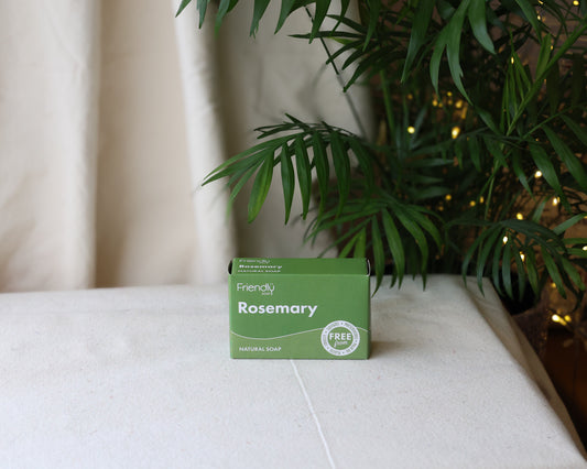 A bar of Rosemary & Nettle Soap placed on a white surface with a green box packaging behind it, featuring a plant and fairy lights in the background.