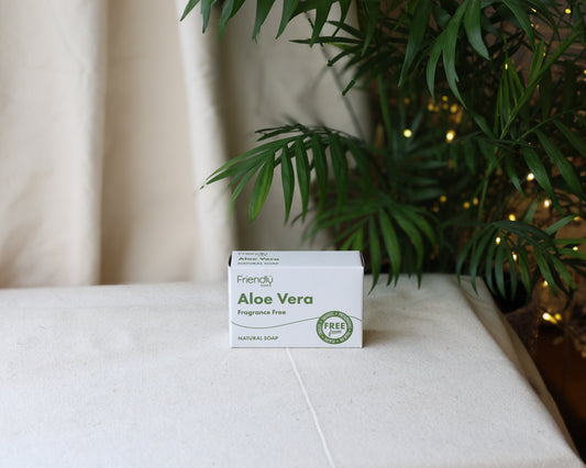 A bar of Aloe Vera fragrance-free soap with packaging that includes the product name and brand, placed on a surface with a plant and lights in the background.