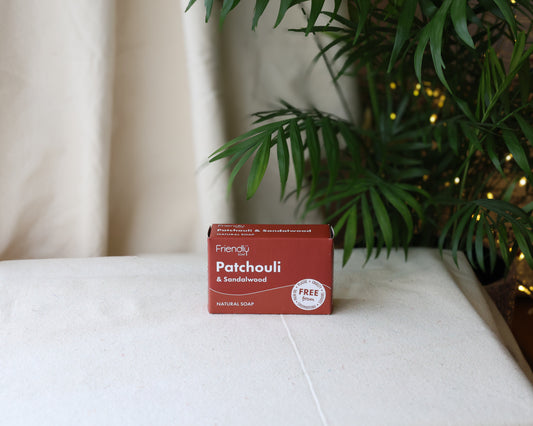 A bar of Patchouli & Sandalwood soap with its packaging, placed on a surface with a plant and Christmas lights in the background.