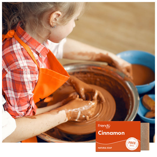 Child in an orange apron working with clay on a pottery wheel, with a Friendly Soap cinnamon bar in the corner.