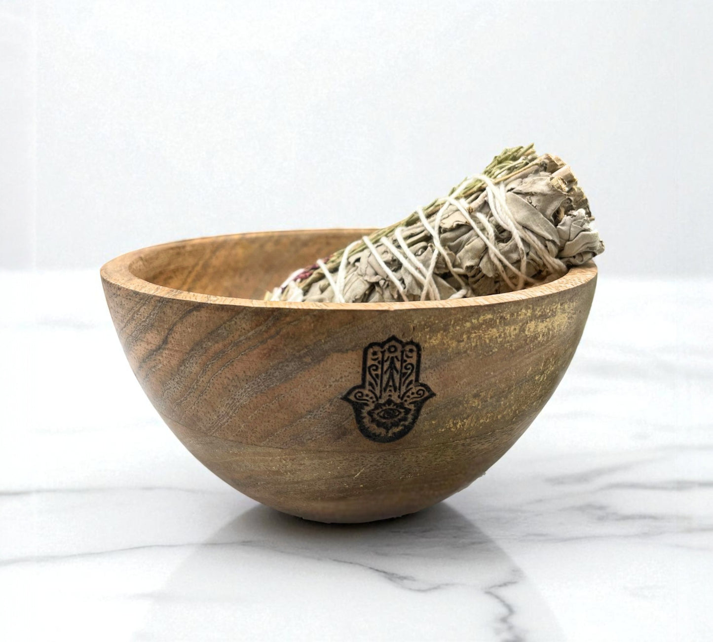Wooden bowl with Hamsa hand design containing sage on a white marble surface