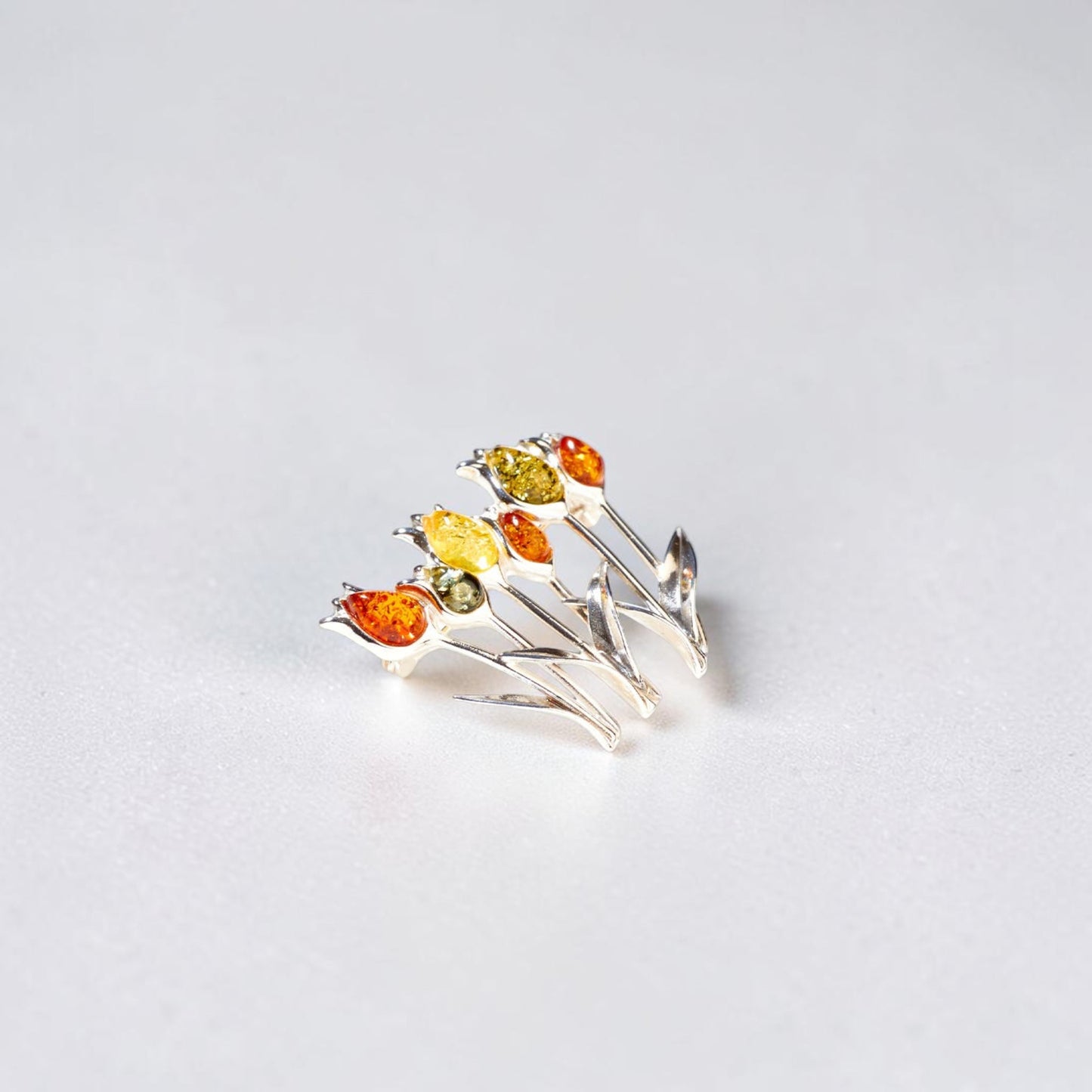 A silver brooch designed as a cluster of tulips, featuring faceted amber gemstones in amber orange, bright yellow, and olive green, set against a plain light grey background.