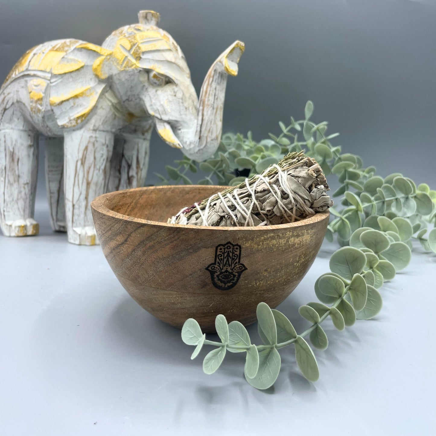 Wooden bowl with sage and a Hamsa hand symbol, next to a decorative elephant and eucalyptus leaves on a light background