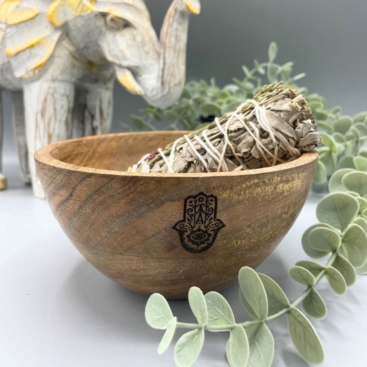Wooden bowl with smudge stick and Hamsa hand symbol on a white surface with greenery.