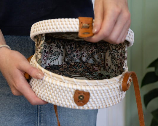 Hands opening a round white rattan Bali bag to show the patterned batik lining, with tan leather strap and clasp