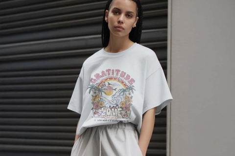  Woman wearing an oversized pale grey t-shirt with a vintage-style Gratitude and Love tropical graphic print featuring palm trees, waves and florals, styled with matching grey wide-leg trousers