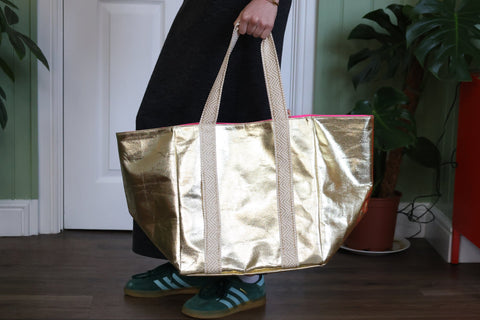  Person carrying an oversized metallic gold tote bag with geometric cream handles and a pink interior lining, styled with a black dress and green Adidas Gazelle trainers in a green hallway