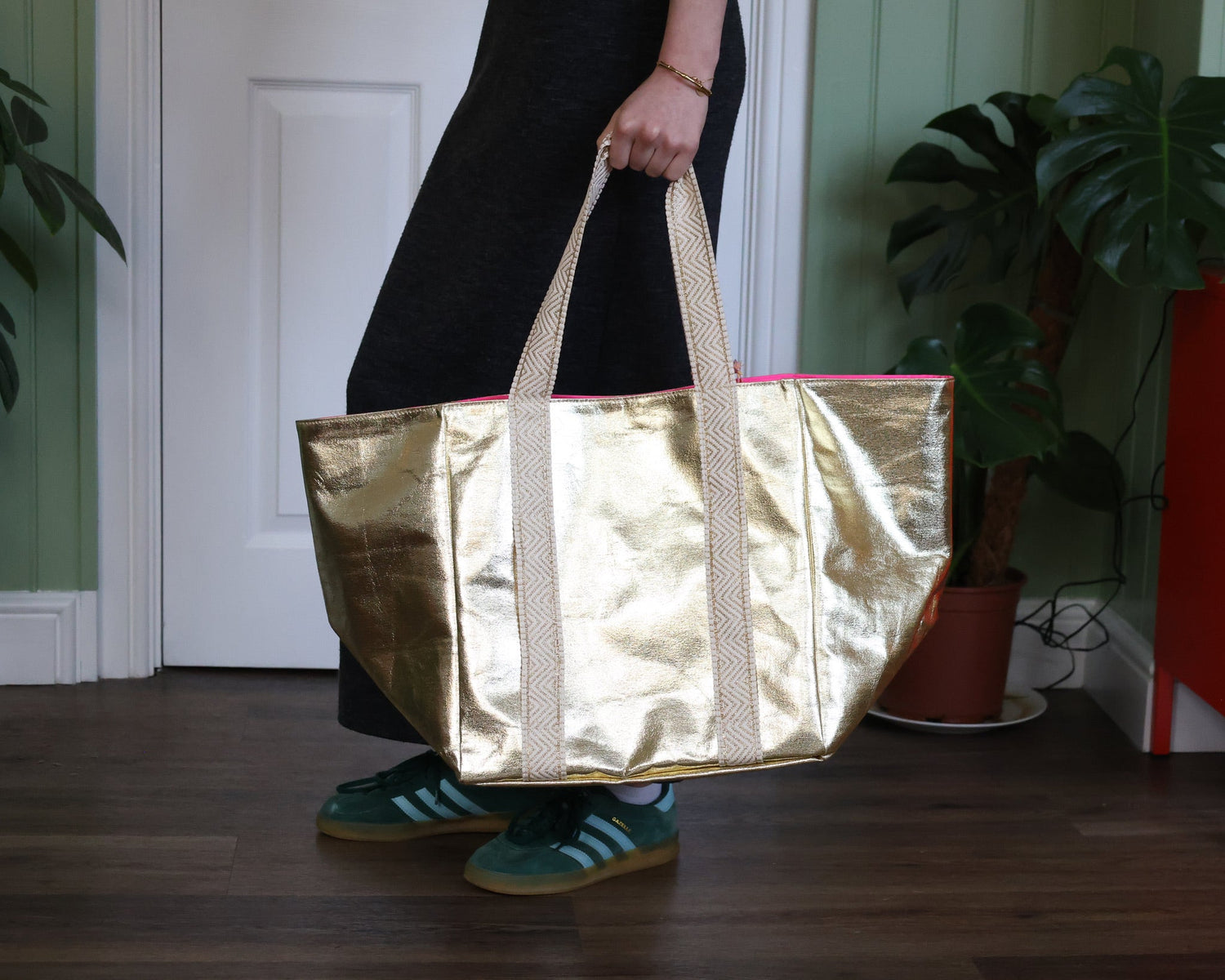 Person carrying an oversized metallic gold tote bag with geometric cream handles and a pink interior lining, styled with a black dress and green Adidas Gazelle trainers in a green hallway