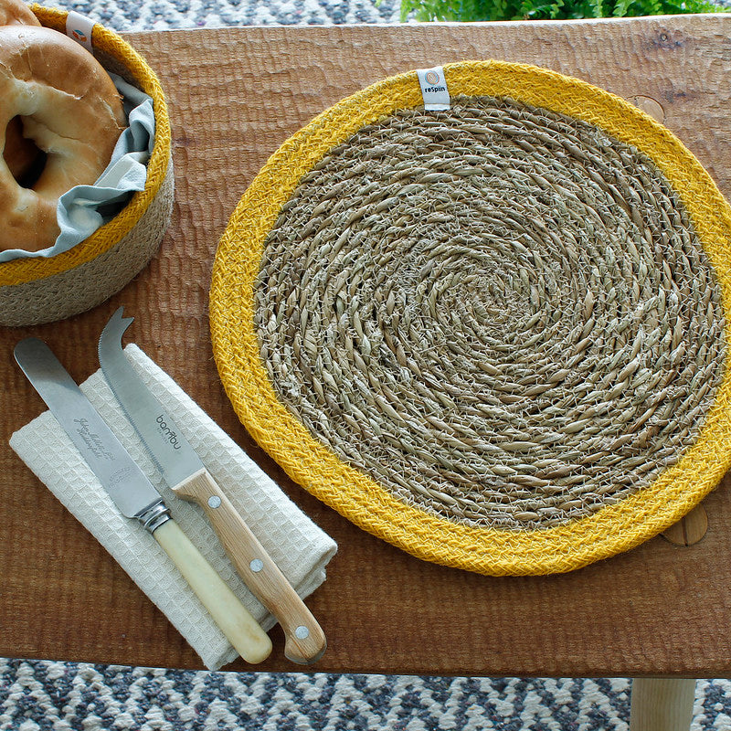 Round woven seagrass placemat with a mustard yellow cotton border, styled on a rustic wooden table with wooden-handled cutlery, a linen napkin and a matching bread basket