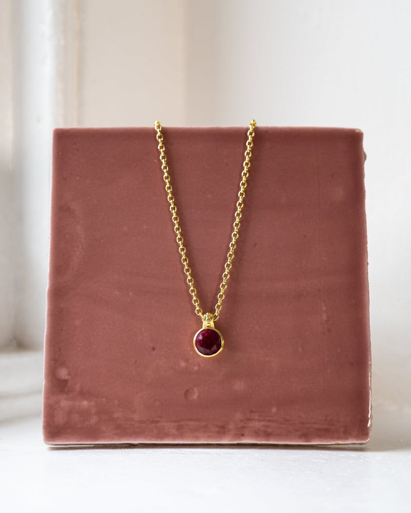 Gold chain necklace with a deep red ruby gemstone pendant in a gold bezel setting, displayed on a dusty pink ceramic tile