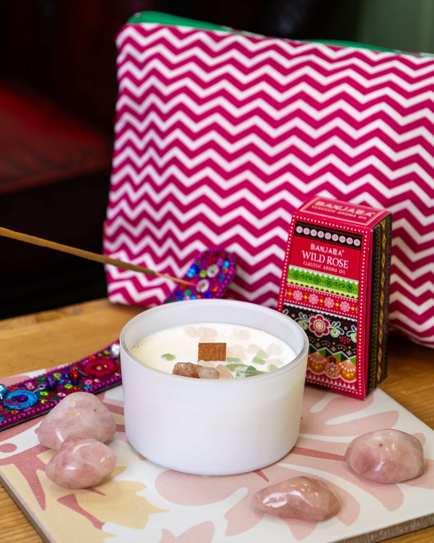 White soy wax crystal candle with a wooden wick surrounded by rose quartz tumblestones, styled with a Banjara Wild Rose aroma oil box, a pink chevron pouch and an incense stick on a wooden surface