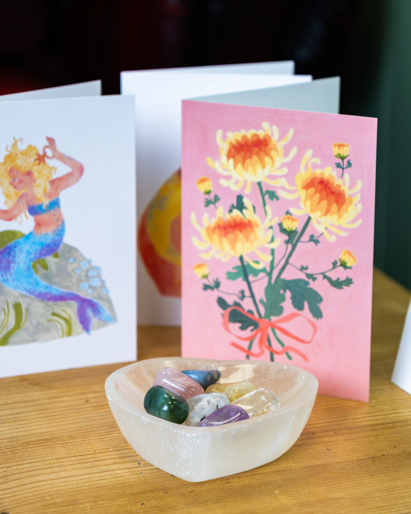 Selenite crystal bowl filled with tumbled gemstones displayed in front of two illustrated greeting cards — a mermaid design and an orange chrysanthemum bouquet on pink — on a wooden surface