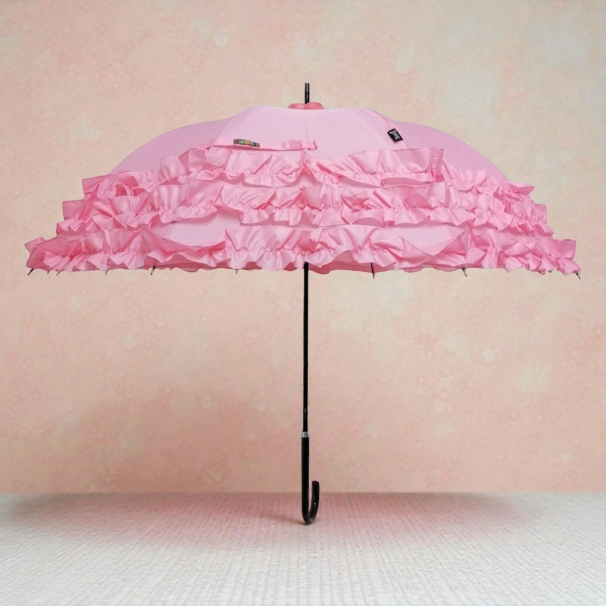 Open pink umbrella with three tiers of satin ruffle trim and a curved black handle, displayed against a soft pink textured background