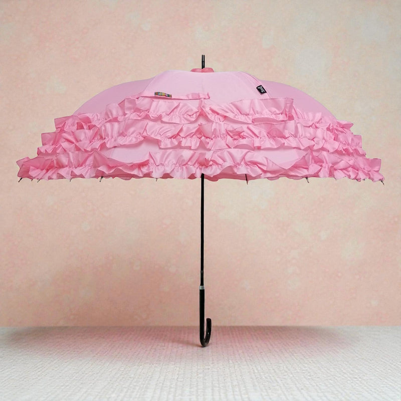 Open pink umbrella with three tiers of satin ruffle trim and a curved black handle, displayed against a soft pink textured background