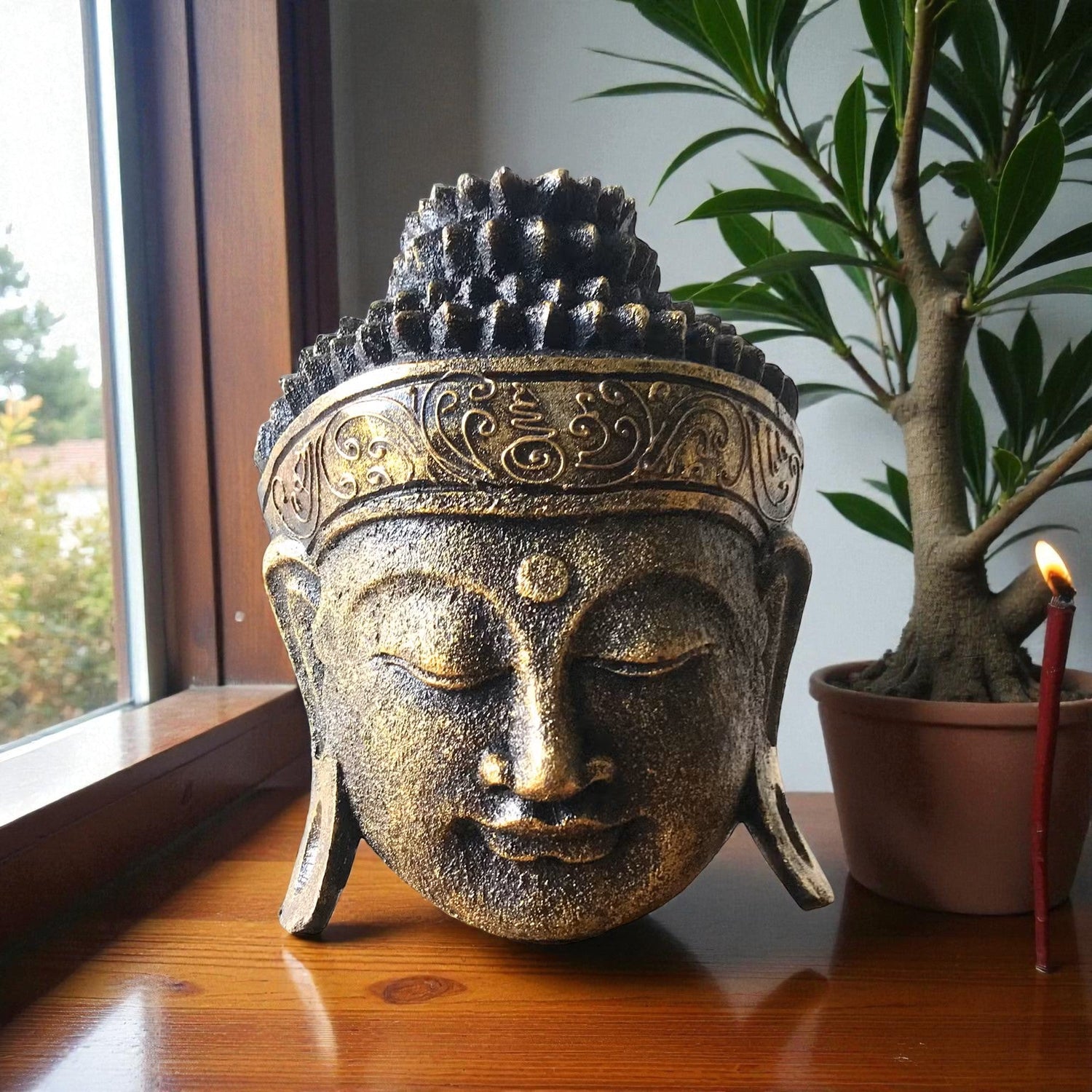 Large dark gold Buddha head sculpture with intricate carved headband detail, displayed on a wooden windowsill beside a potted bonsai tree and a lit red candle