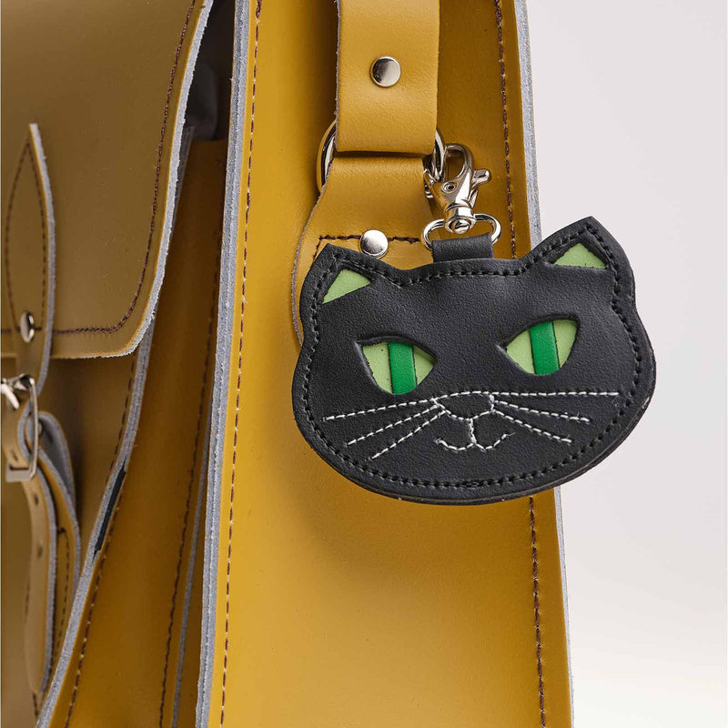Black leather cat face bag charm with green striped eyes and stitched whisker detail, clipped onto the strap of a mustard yellow leather satchel