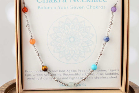  Little Gems Jewels chakra necklace with seven coloured gemstone beads on a silver chain, presented in a kraft gift box, displayed on a wooden dish with matching tumblestones including amethyst, turquoise and carnelian