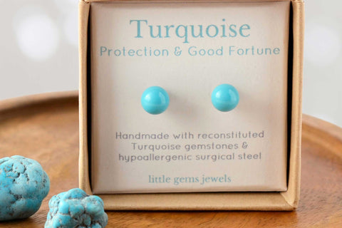  Little Gems Jewels turquoise gemstone stud earrings presented in a kraft gift box with Protection & Good Fortune text, displayed on a wooden dish with raw turquoise stones