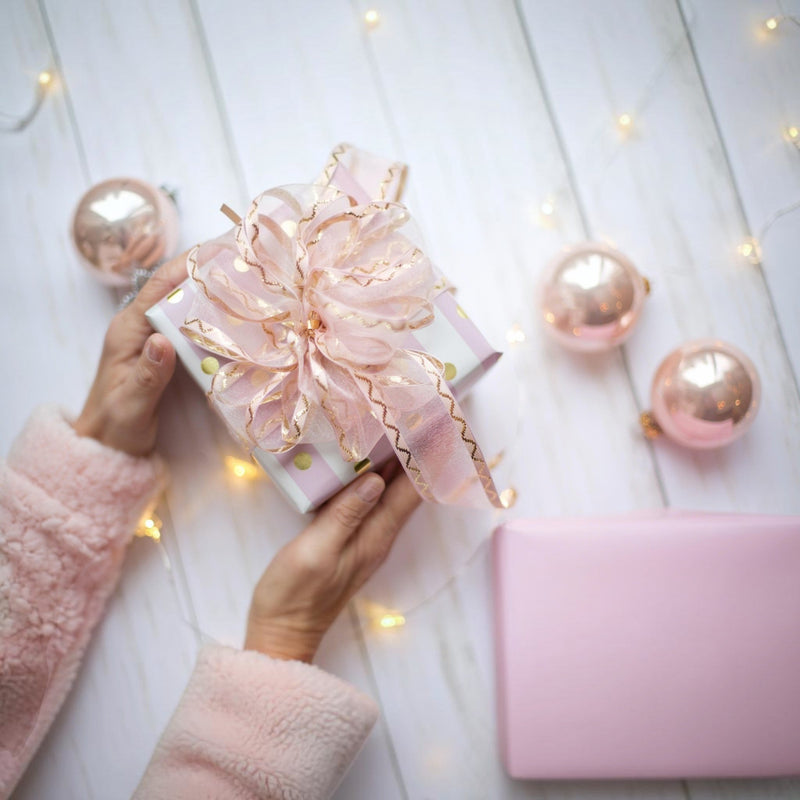 Hands holding a pastel pink gift box with a gold‑trimmed ribbon, surrounded by blush ornaments and fairy lights — Apricot Moon Gifts.