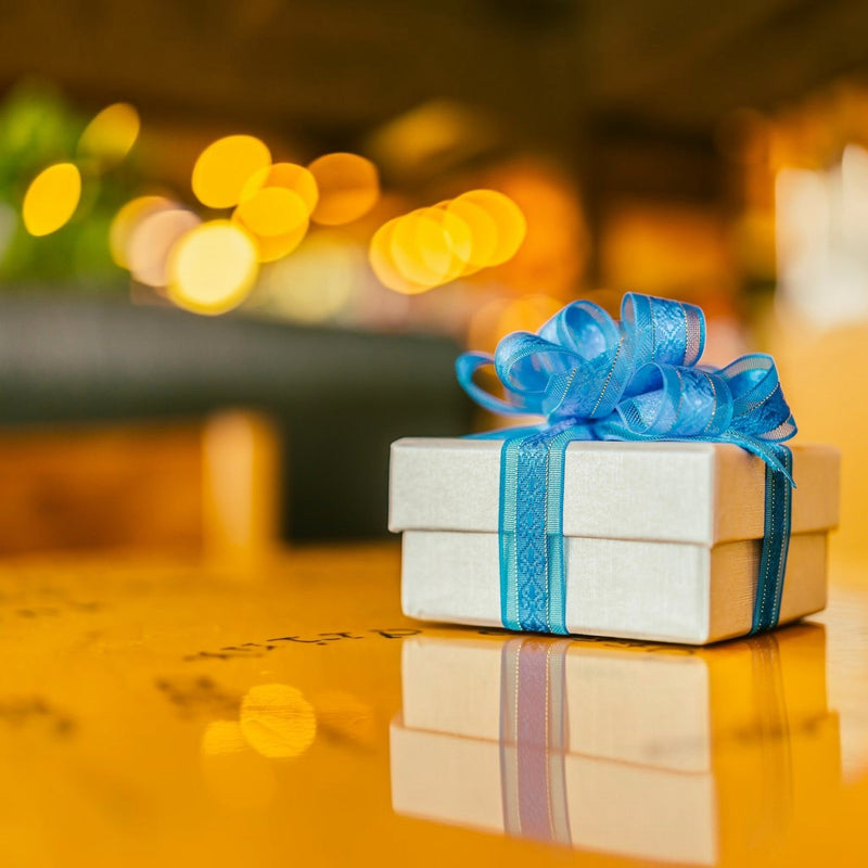 Small gift box wrapped with a blue satin ribbon on a golden table, surrounded by warm glowing lights — Apricot Moon Gifts Under £20.