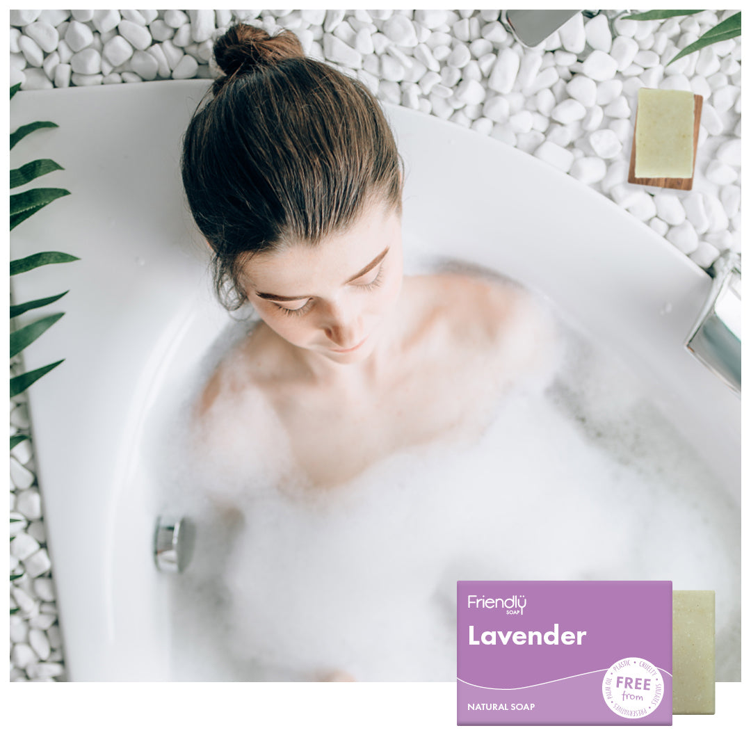 Woman relaxing in a deep bubble bath surrounded by white pebbles and tropical leaves, with a Friendly Soap Lavender natural soap bar and packaging shown in the corner