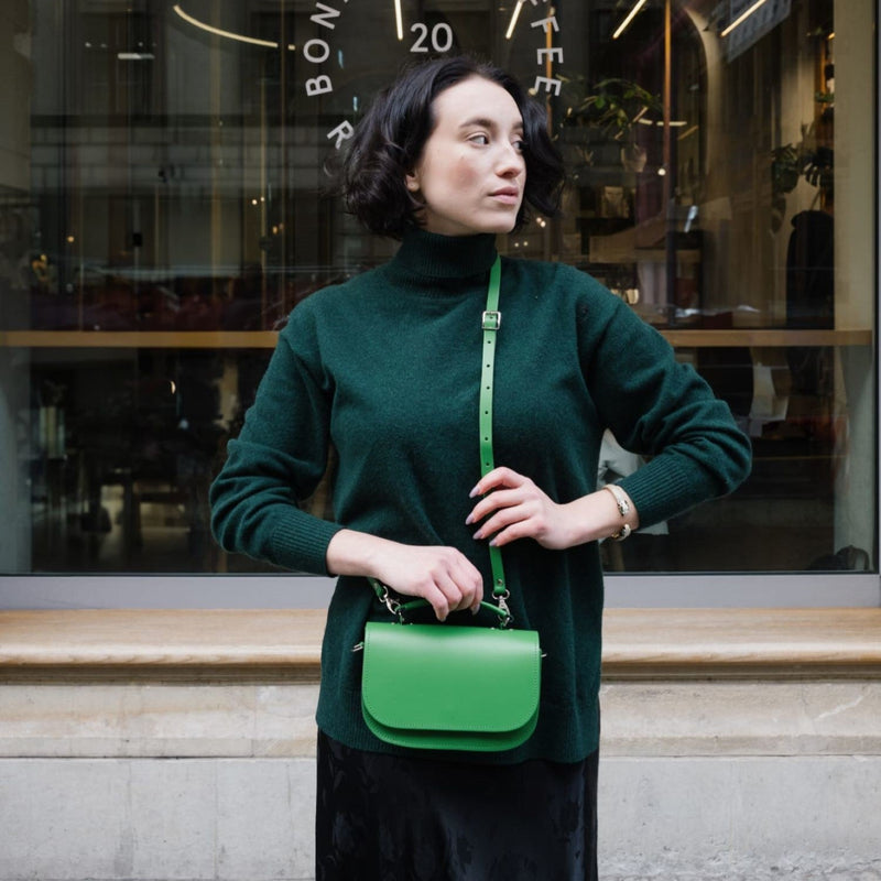 Woman wearing a dark green turtleneck jumper and black velvet skirt carrying a bright green leather crossbody saddle bag on an adjustable strap, photographed outside a coffee shop