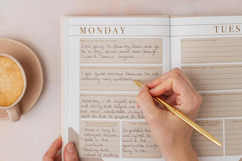  Person writing in an open mindfulness journal with daily intention and gratitude prompts, styled with a flat white coffee and croissant on a pink surface