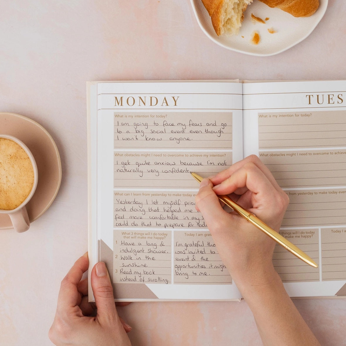 Person writing in an open mindfulness journal with daily intention and gratitude prompts, styled with a flat white coffee and croissant on a pink surface