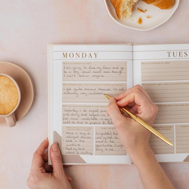 Person writing in an open mindfulness journal with daily intention and gratitude prompts, styled with a flat white coffee and croissant on a pink surface