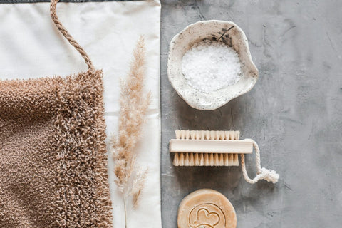  Natural bath and body essentials arranged on a textured grey surface — handmade soap with heart motif, wooden brush, mineral bath salts, soft towel and dried grass stems, evoking calm, sustainable self‑care.