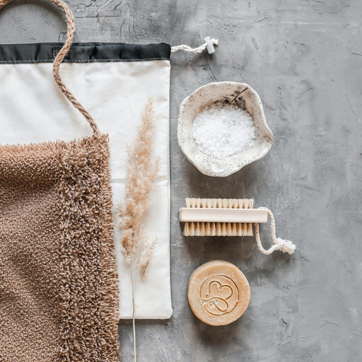 Natural bath and body essentials arranged on a textured grey surface — handmade soap with heart motif, wooden brush, mineral bath salts, soft towel and dried grass stems, evoking calm, sustainable self‑care.
