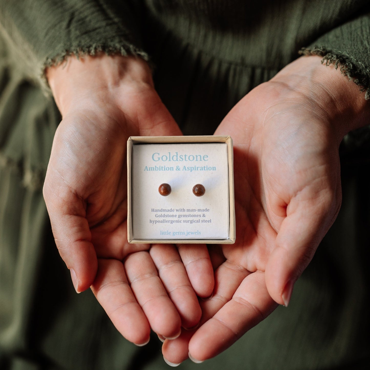 Hands holding a box of Goldstone earrings against a dark background