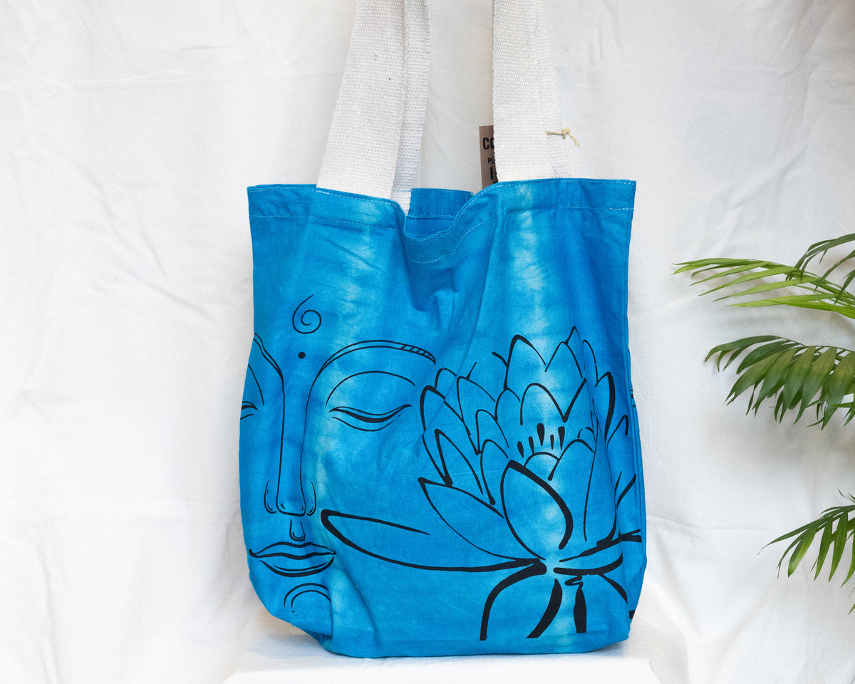 A bright blue fabric tote bag with black line art of a Buddha face and a lotus flower, hanging against a white fabric backdrop with a green plant leaf visible on the right.