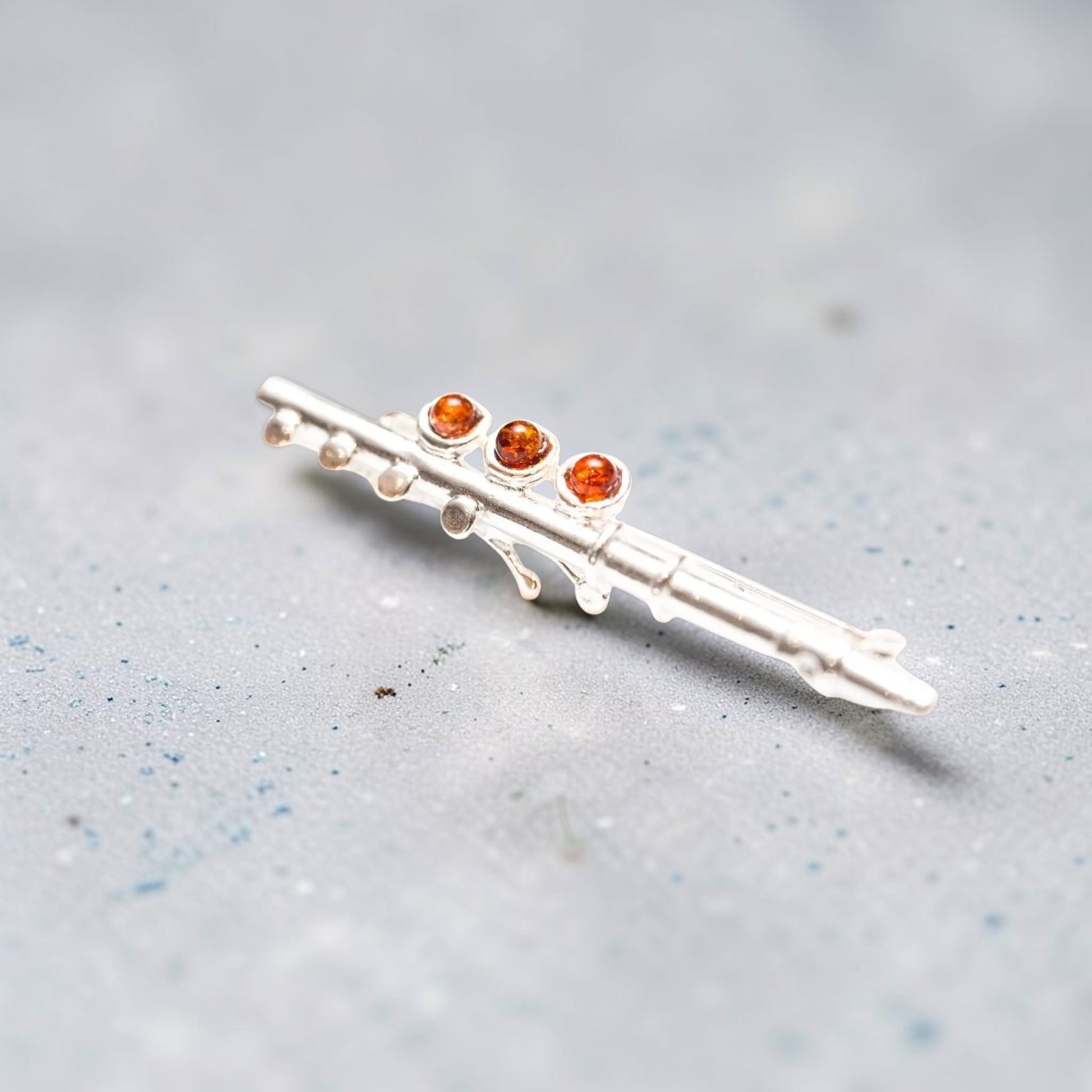 A silver-toned flute-shaped brooch with three small round amber-orange gemstones, resting on a textured light grey surface.