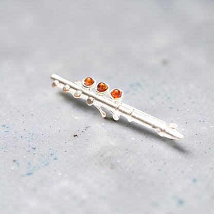 A silver-toned flute-shaped brooch with three small round amber-orange gemstones, resting on a textured light grey surface.