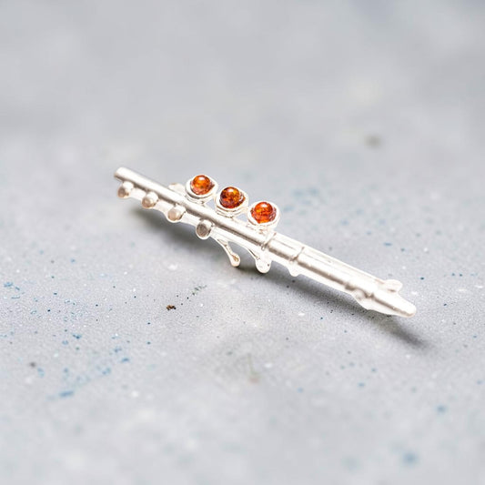 A silver-toned flute-shaped brooch with three small round amber-orange gemstones, resting on a textured light grey surface.