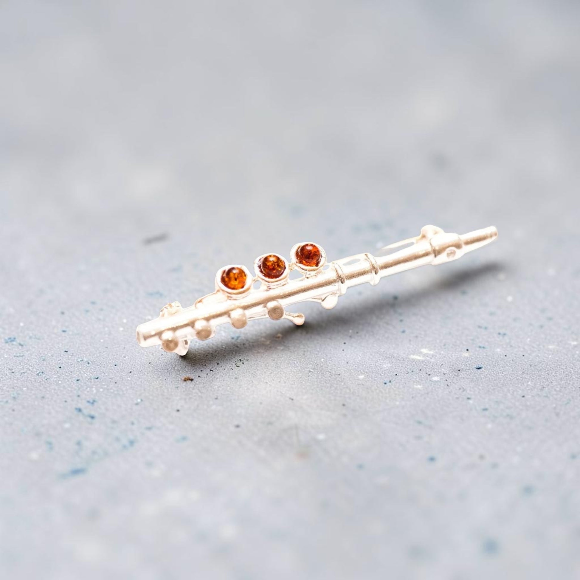A miniature gold flute-shaped brooch with three round amber-orange gemstones, resting on a textured gray surface.