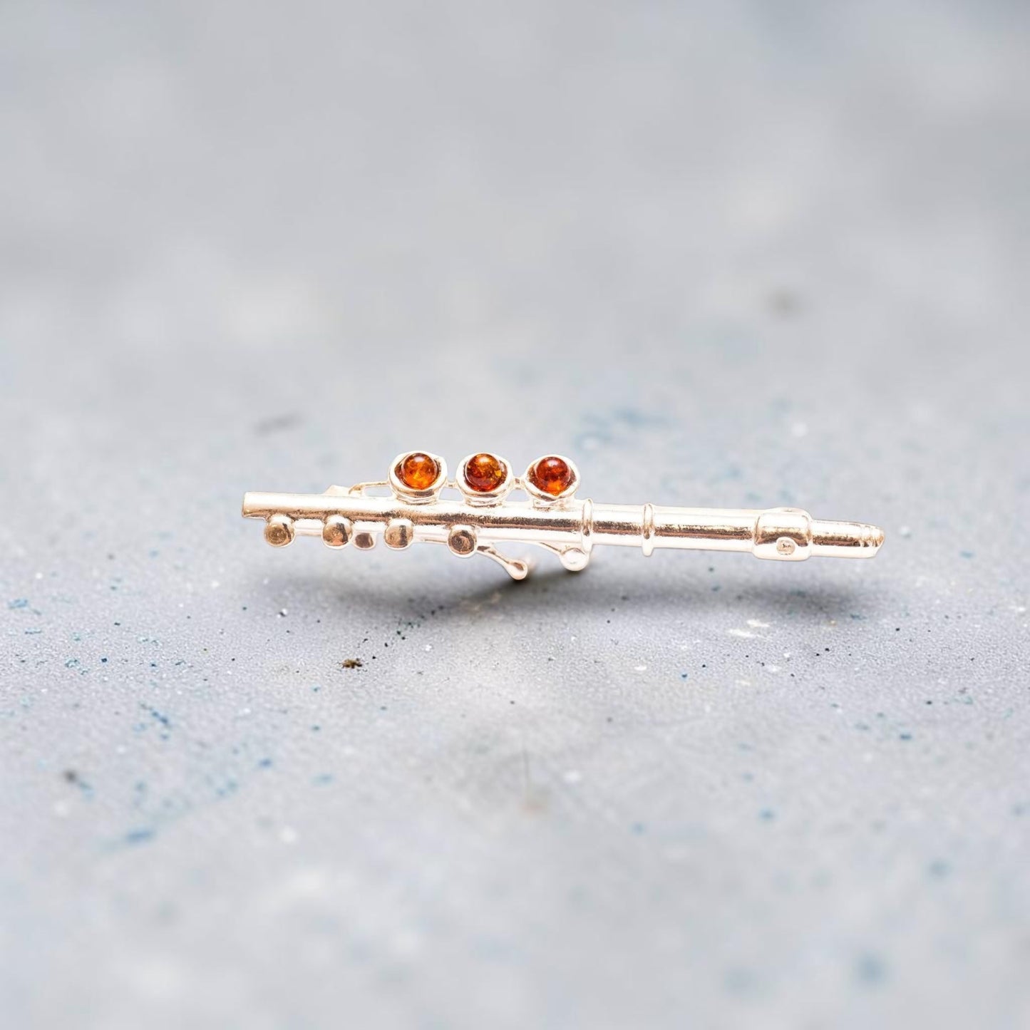 A miniature gold-tone flute-shaped pin or brooch with three small round amber-orange gemstones, photographed on a soft gray textured background.