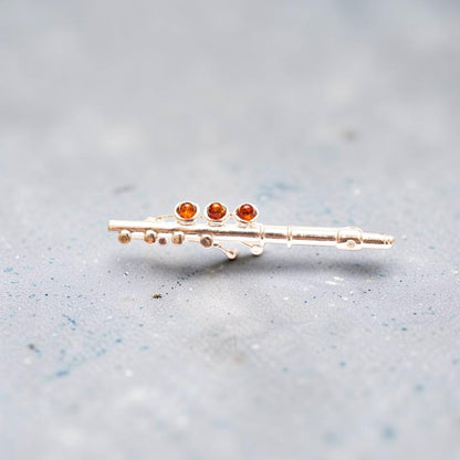 A miniature gold-tone flute-shaped pin or brooch with three small round amber-orange gemstones, photographed on a soft gray textured background.