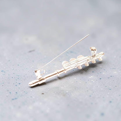 A silver flute-shaped brooch with a thin pin back, displayed on a textured gray surface.