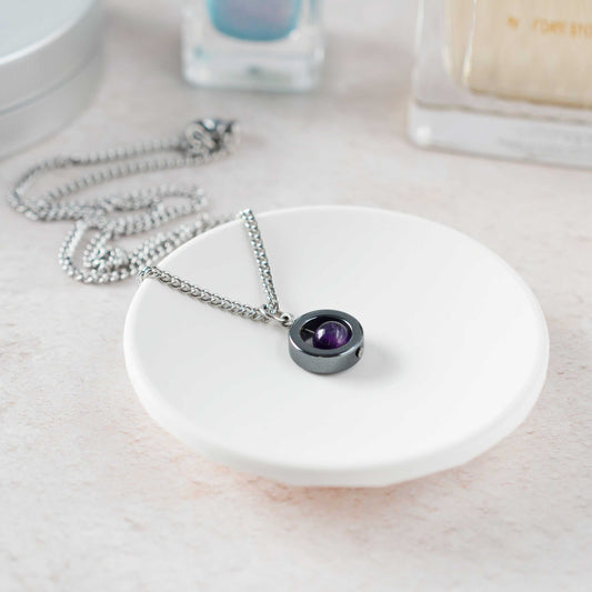Necklace with a purple gemstone pendant on a white dish, with a blurred background of bottles.