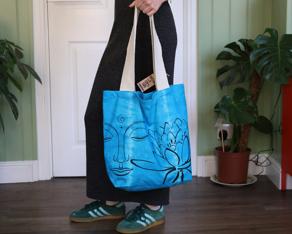 A person wearing a long black dress and green sneakers holds a bright blue canvas tote bag with a black line art design of a face and lotus flower. The setting is indoors with a white door, green paneled walls, and potted plants.