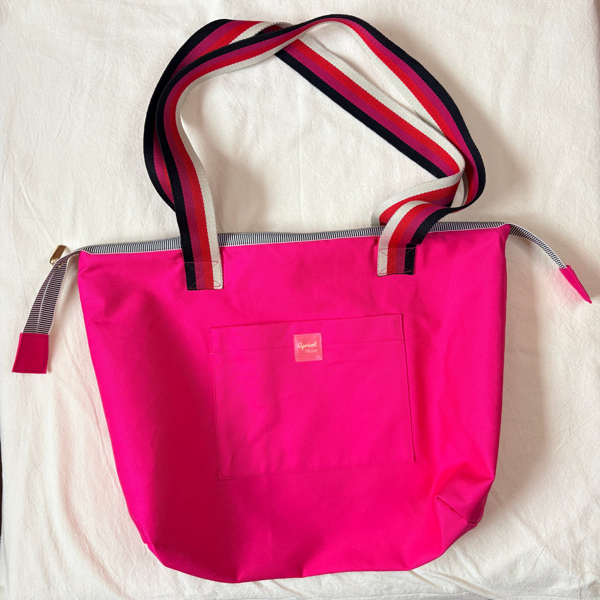 Berry Riot bright pink shoulder tote laid flat on ivory fabric, with pink, red, cream and black striped handles and a front pocket with a pink Apricot Moon label, handmade in Dorset by Apricot Moon