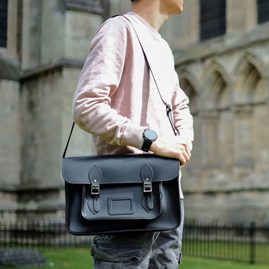 Person holding a black leather satchel bag in front of a stone building