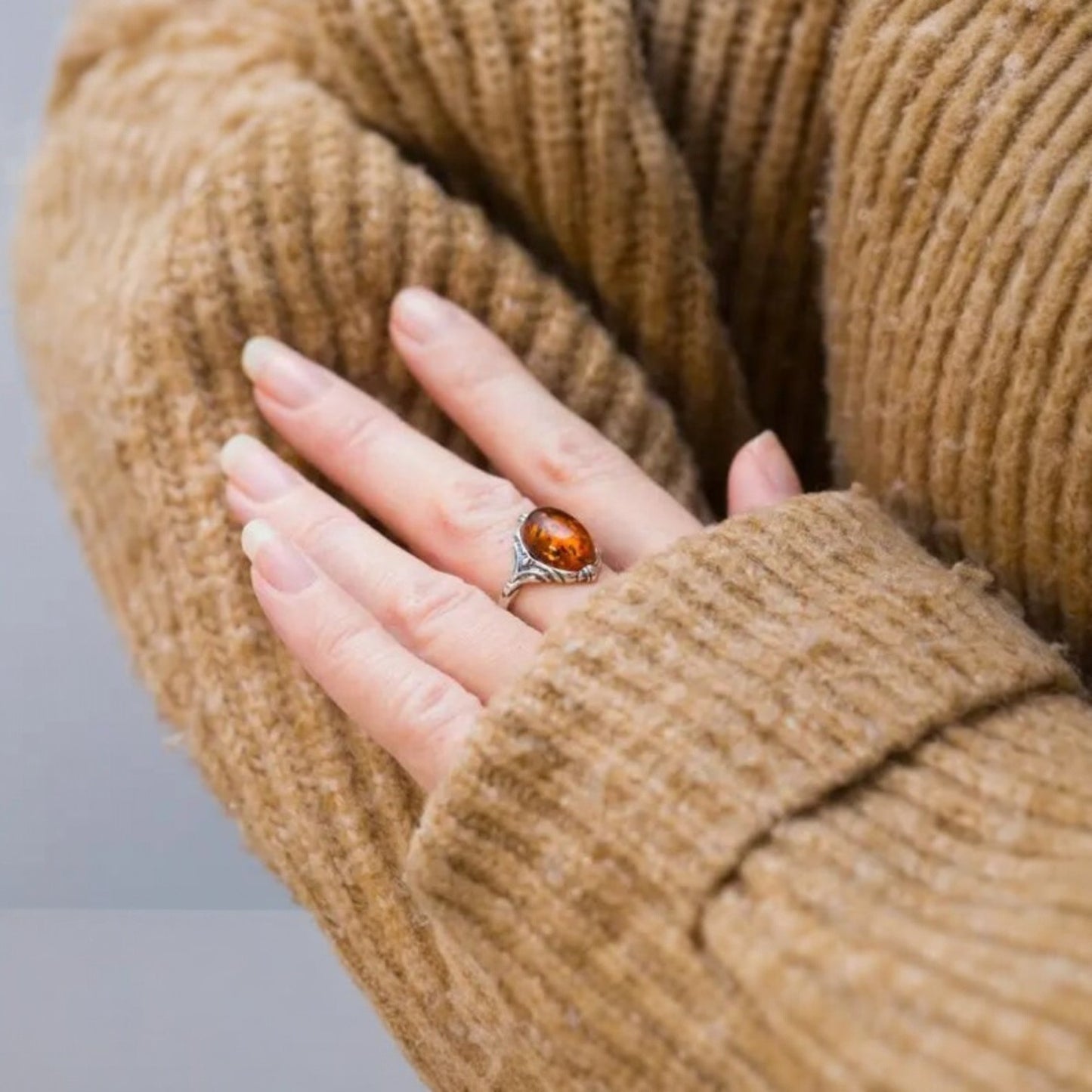 Hand wearing a ring with an amber stone, wrapped in a brown textured sweater.