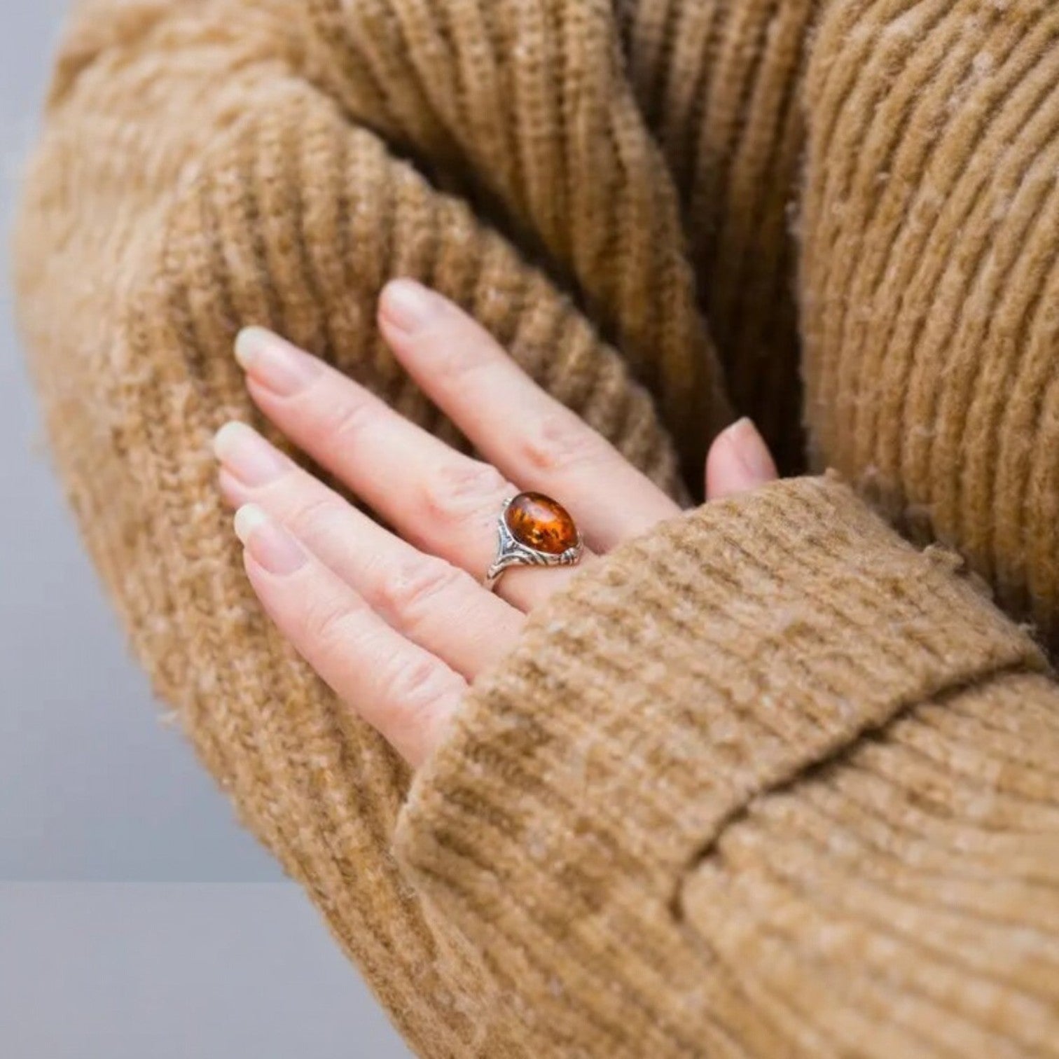 Hand wearing a ring with an amber stone, wrapped in a brown textured sweater.