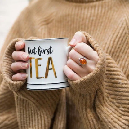 Person holding a mug with 'but first tea' text, wearing a brown sweater.
