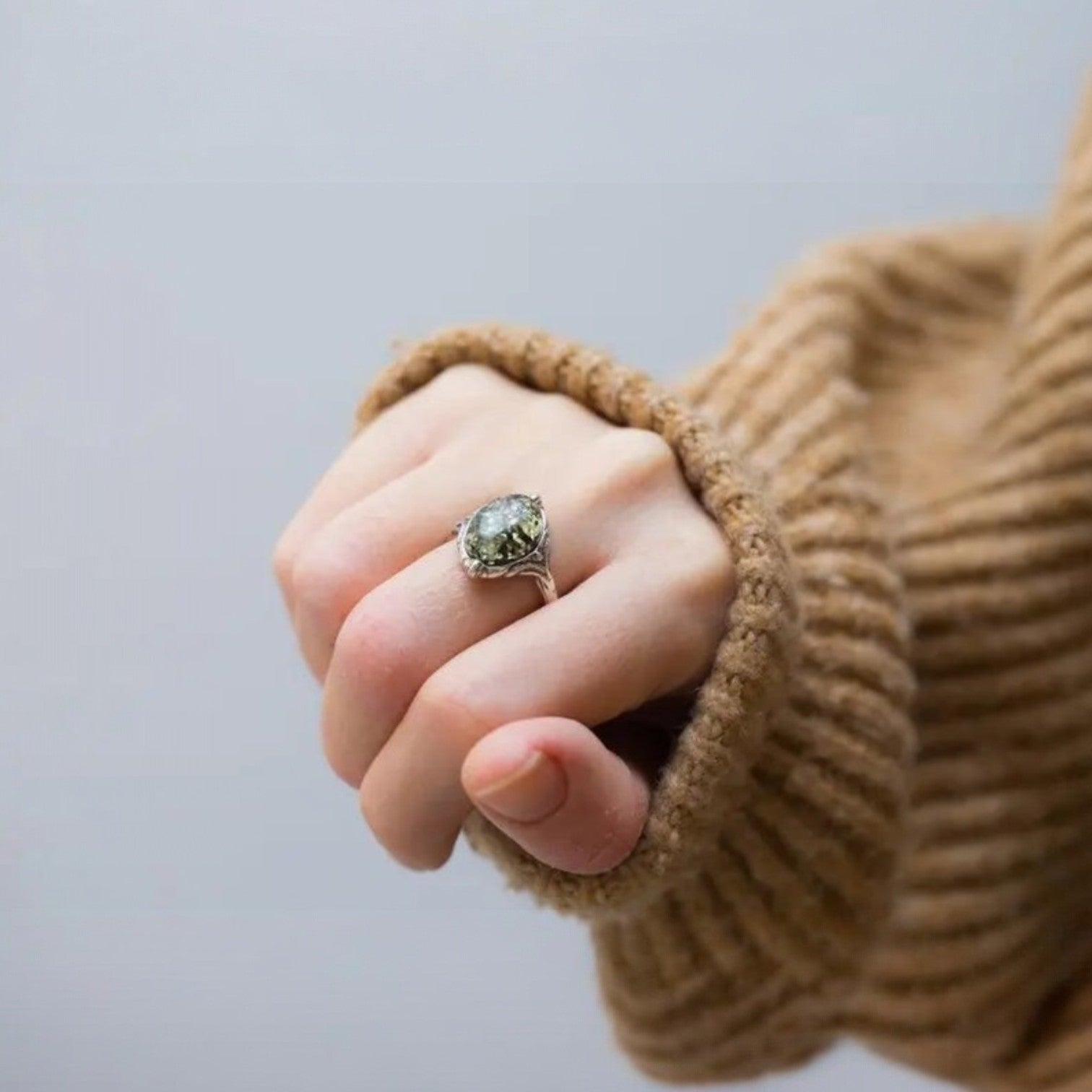 Hand wearing a ring with a green gemstone, wearing a brown sweater.