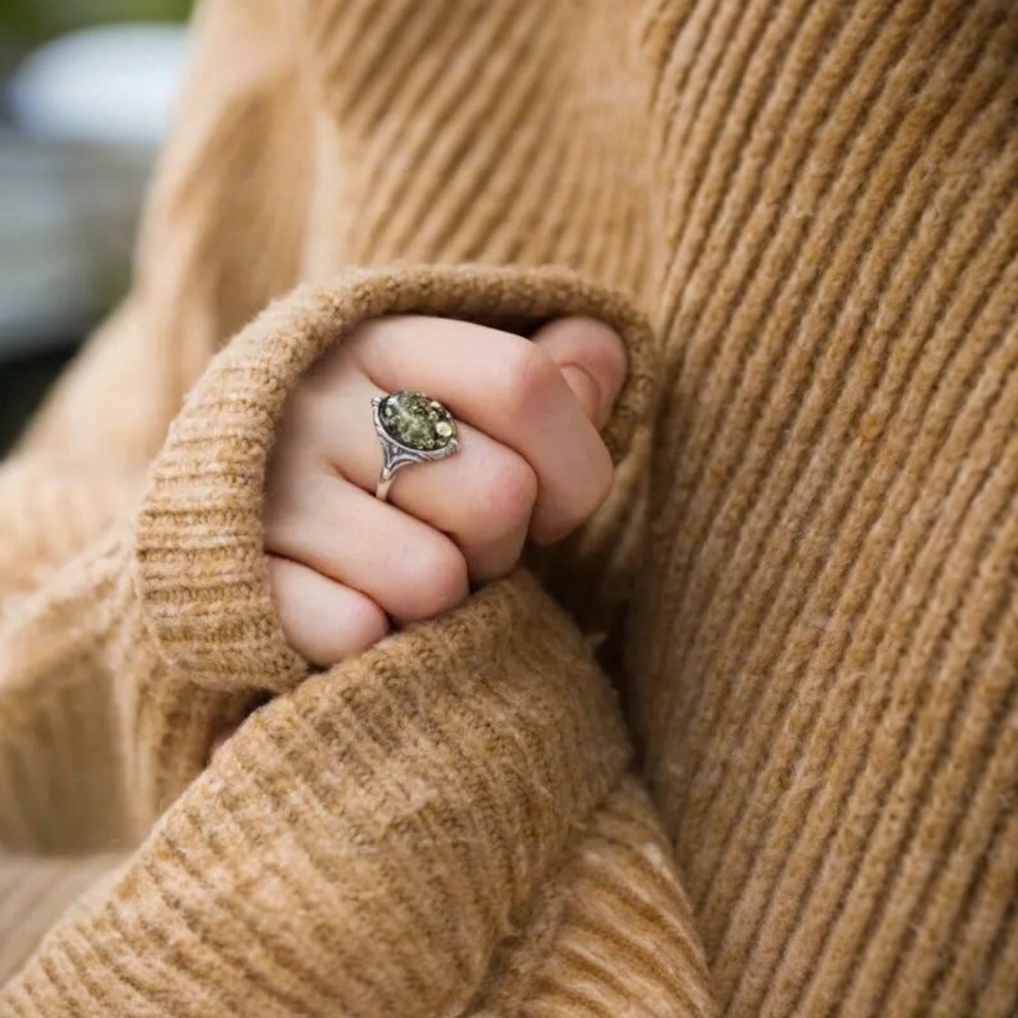 Close-up of a hand wearing a ring with a green gemstone, wrapped in a beige sweater.