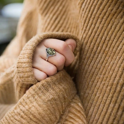 Close-up of a hand wearing a ring with a green gemstone, wrapped in a beige sweater.